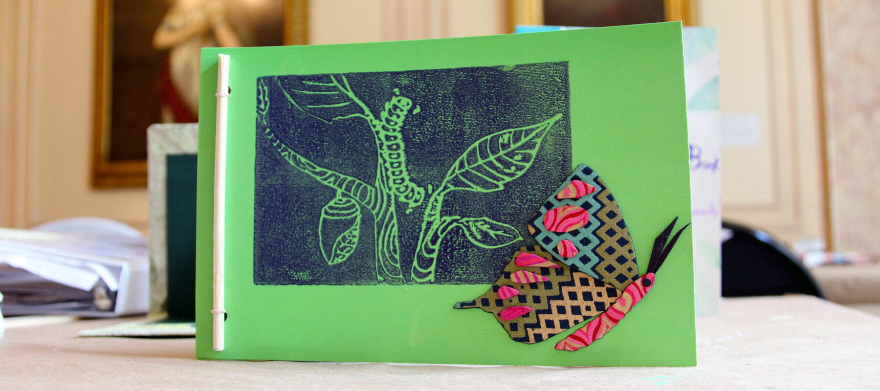 A handmade card displayed on a table with a green cover featuring a black and white textured print of a tree and leaves, and a colorful cut-out butterfly with pink, blue, and yellow patterns. Blurred paintings are in the background.