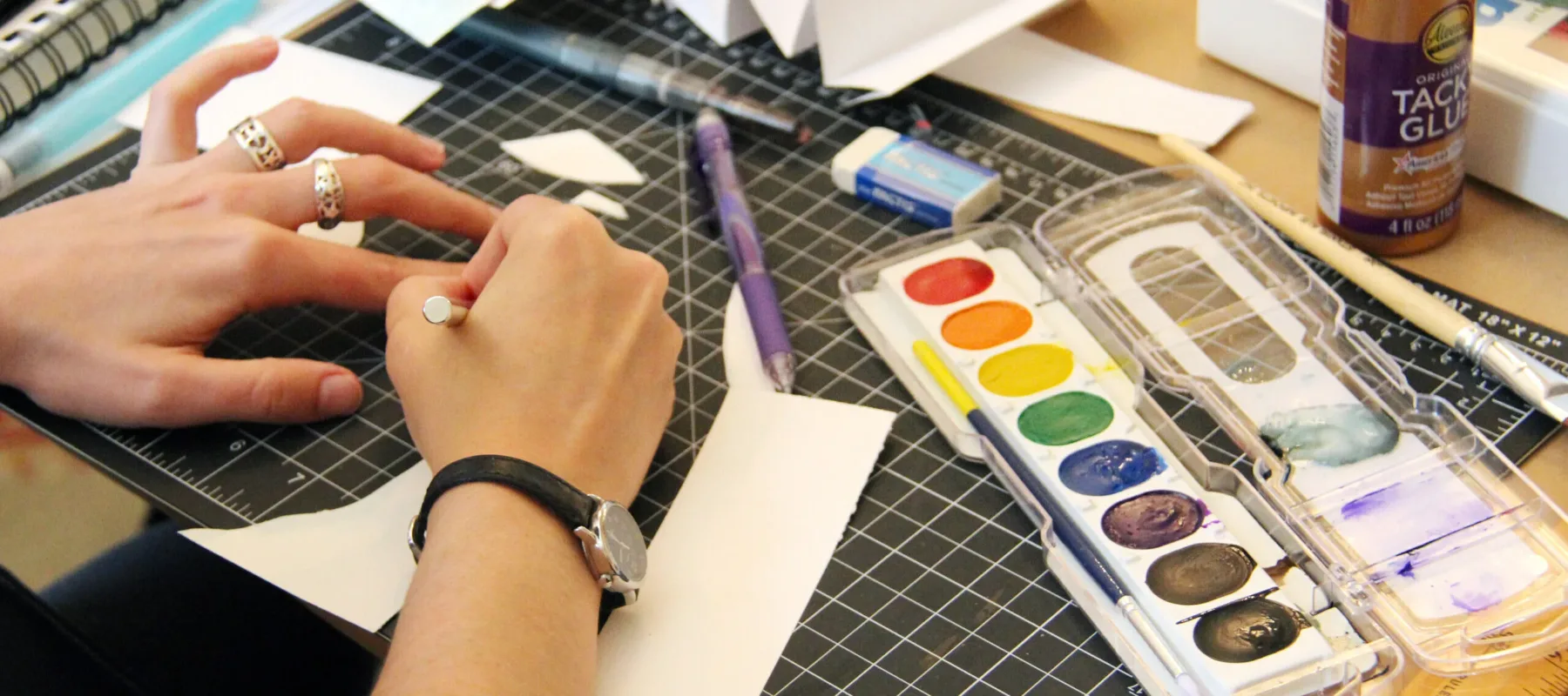 A person with rings on their fingers is drawing on a piece of paper with a pencil. Various art supplies, including a watercolor paint set, an eraser, tacky glue, and a folding paper project are on the table. A cutting mat is used as the workspace surface.