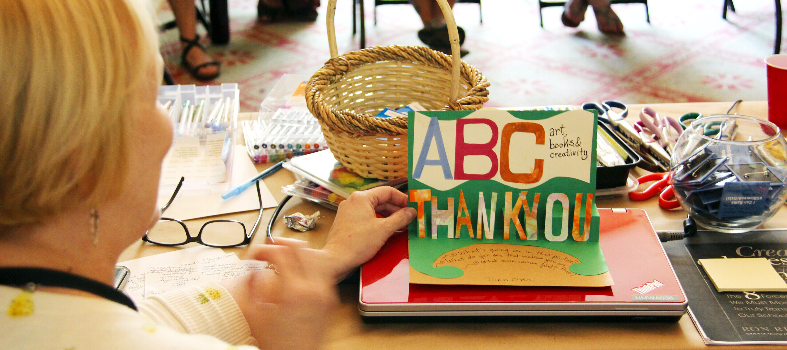 A person seated at a table, holding up a handmade card that says "ABC Thank You" in colorful letters. The table is covered with various craft supplies, including markers, scissors, a basket, and papers. Other people are seated around the room in the background.