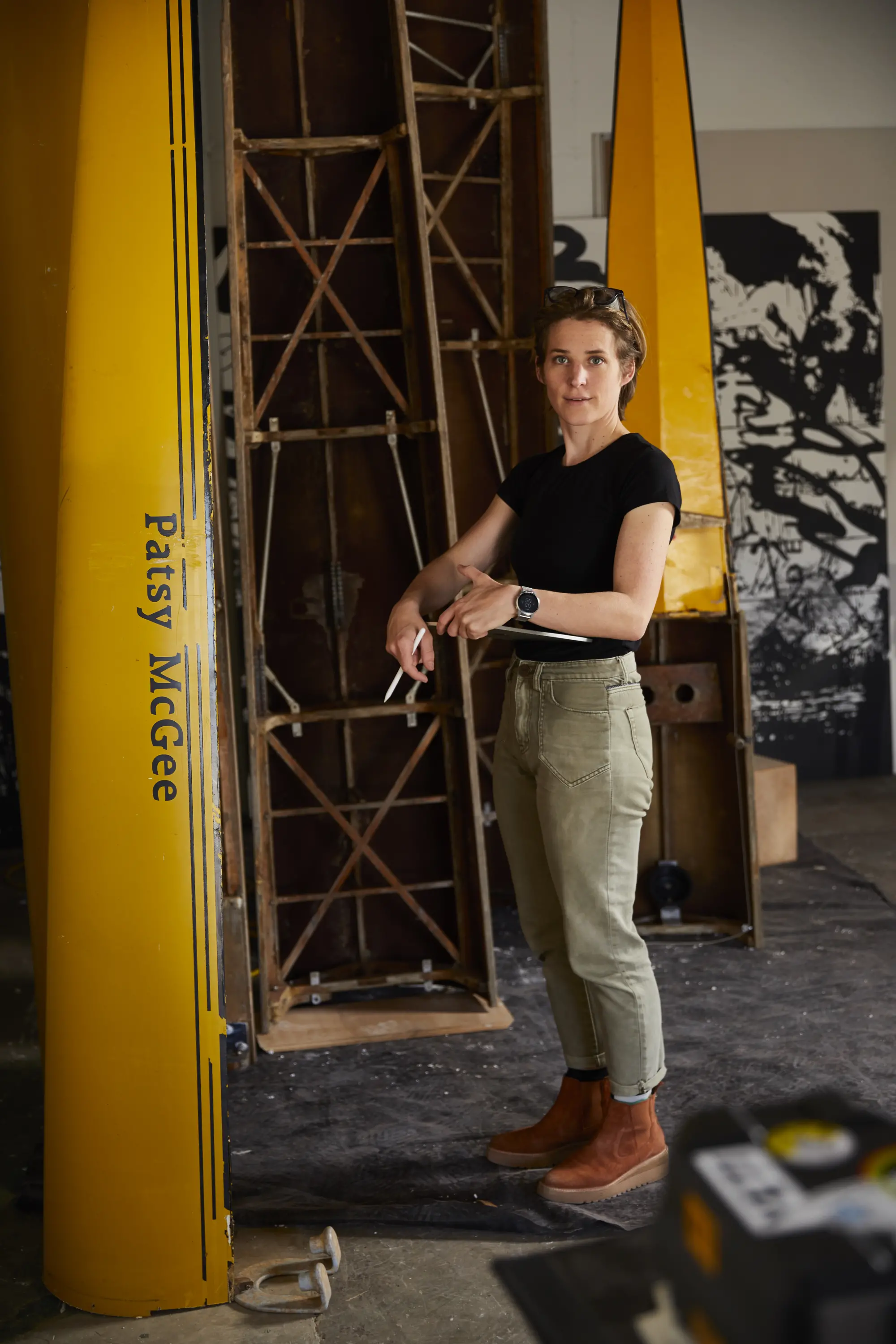 A person standing in a workshop, dressed in a black t-shirt, light green pants, brown boots, and a watch. They are holding tools and standing near a yellow structure with "Patsy McGee" written on it, and part of an unidentified yellow structure are visible in the background.