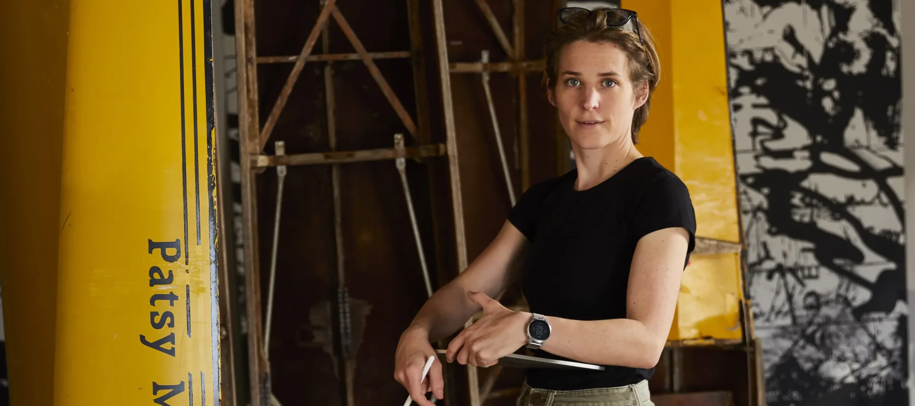 A person standing in a workshop, dressed in a black t-shirt, light green pants, brown boots, and a watch. They are holding tools and standing near a yellow structure with "Patsy McGee" written on it, and part of an unidentified yellow structure are visible in the background.