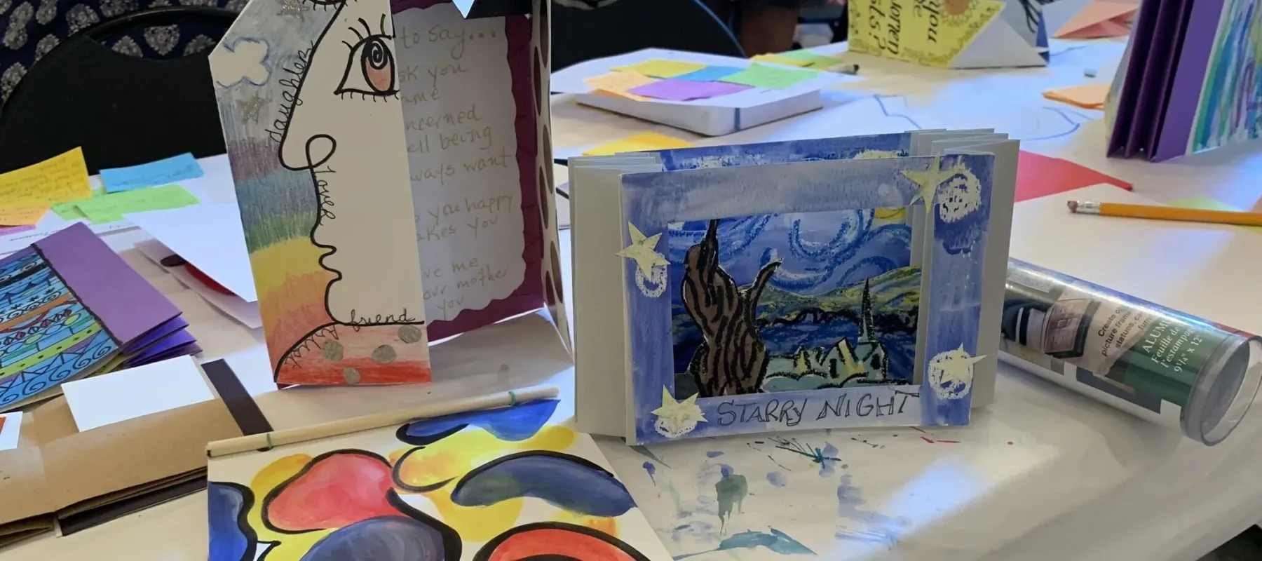 A table is covered with various artistic projects, including a painting inspired by Van Gogh's "Starry Night," abstract art, and a sculpture featuring a face profile. Art supplies like pencils, markers, and paper are scattered on the table. Colorful sticky notes are also visible.