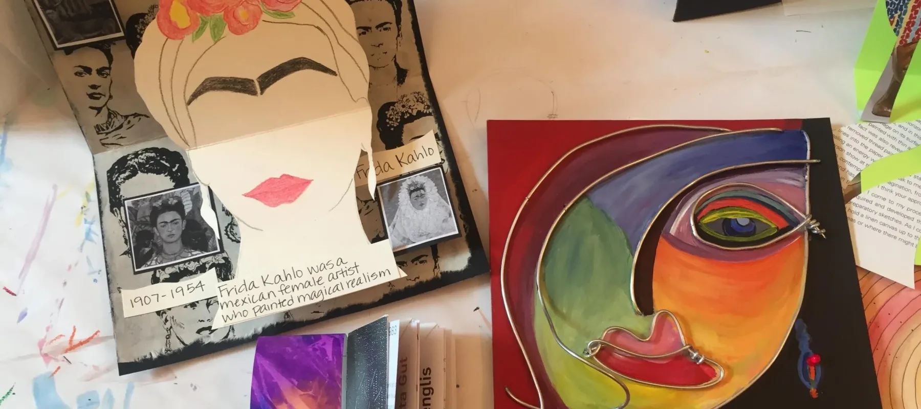 An art table displays two colorful artworks. The piece on the left is a collage with images and quotes about Frida Kahlo, featuring her portrait. The artwork on the right is a vibrant abstract painting of a face. Various art materials and scraps surround the pieces.