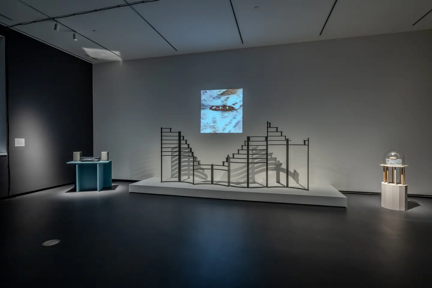 An art exhibition features a minimalist room with a staircase-like sculpture on a white platform, a spherical object on a stand to the right, and a blue table with objects to the left. A video is projected onto the wall behind the sculpture.