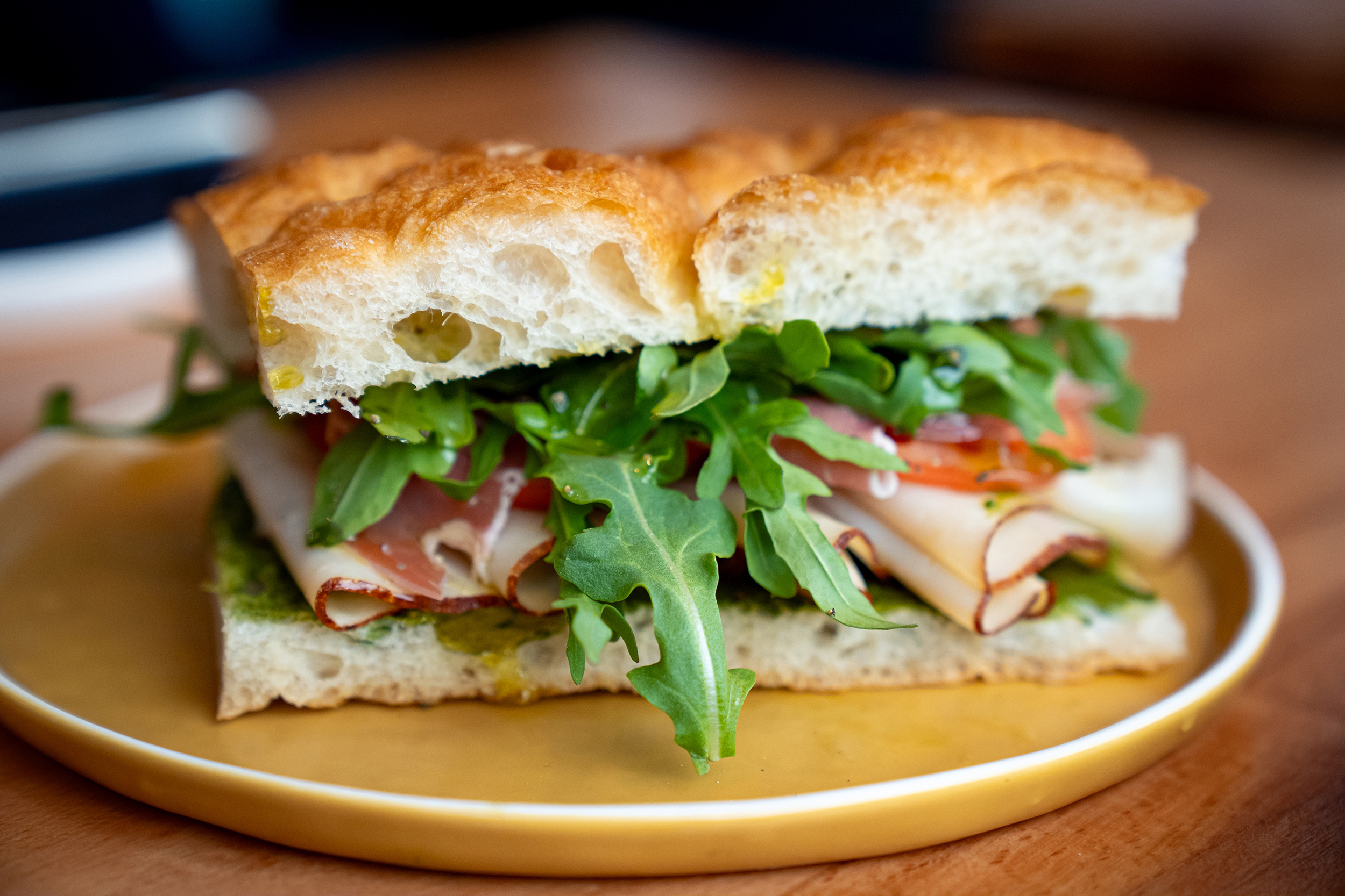 A close-up photo of a sandwich sitting on a yellow plate on a wood table. The sandwich has focaccia bread, arugula, tomato, prosciutto, turkey, and pesto.