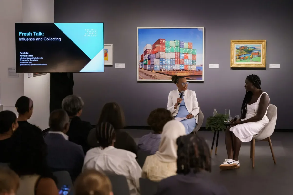 Two women with dark colored skin sit in chairs and talk to each other with microphones.