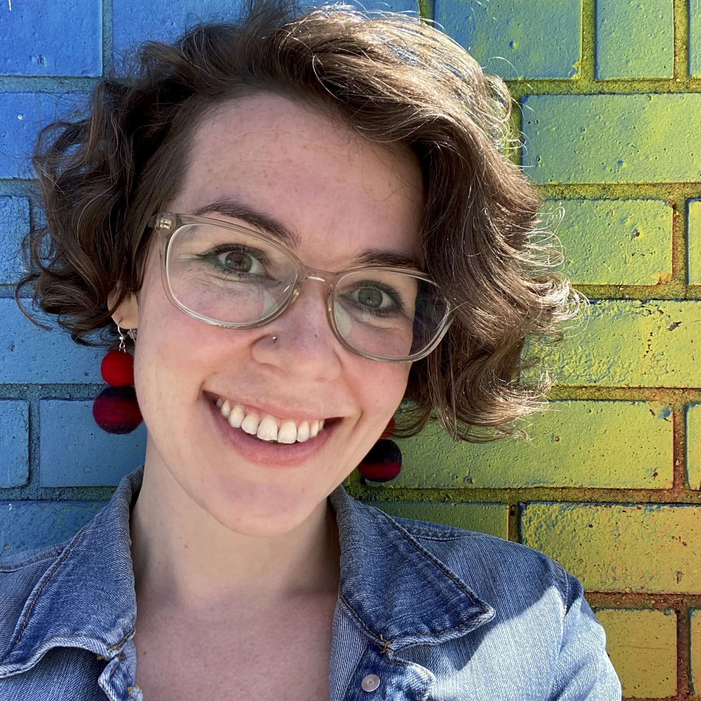 A light-skinned women with ear-length brown hair smiles at the camera in front of a colorful brick wall. She wears wide-rim glasses and red earrings.