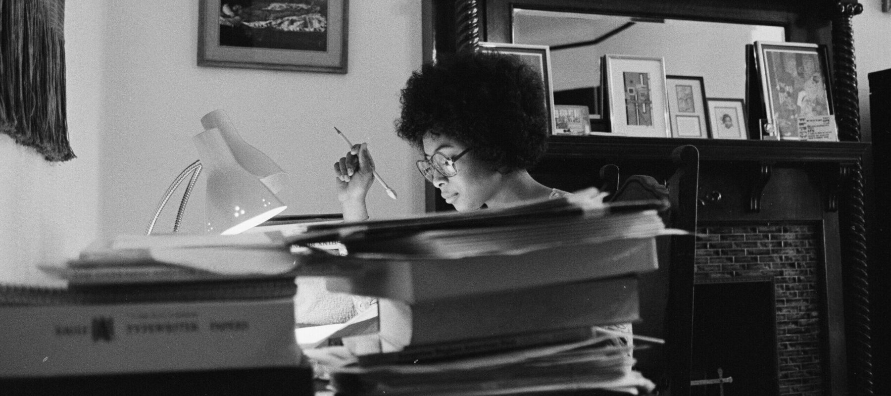Black and white photo of a person with curly hair, wearing glasses, sitting at a desk with a lamp, writing or studying. The desk is cluttered with books and papers. In the background, framed photos and artwork are visible on a mantel.