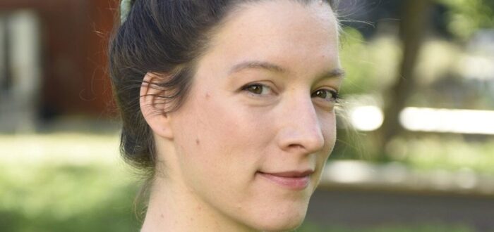 A close-up portrait of a light-skinned woman with brown hair worn in a bun. She turns toward the camera from the side with a cheeky smile.