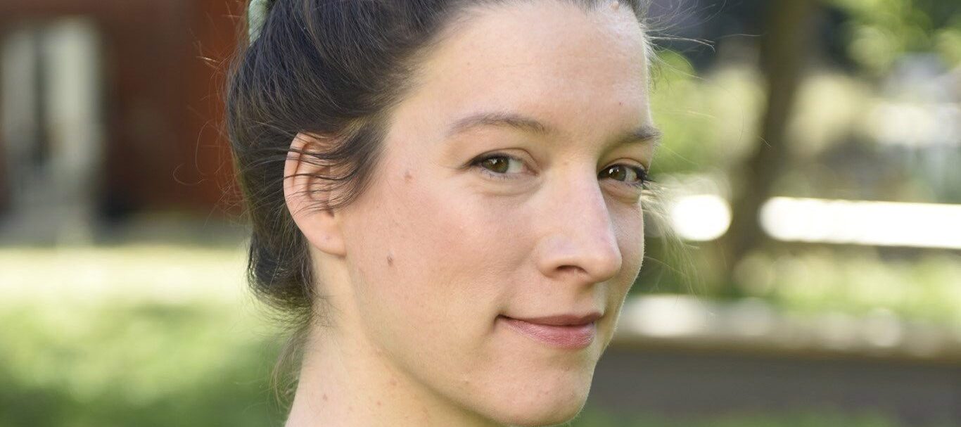 A close-up portrait of a light-skinned woman with brown hair worn in a bun. She turns toward the camera from the side with a cheeky smile.