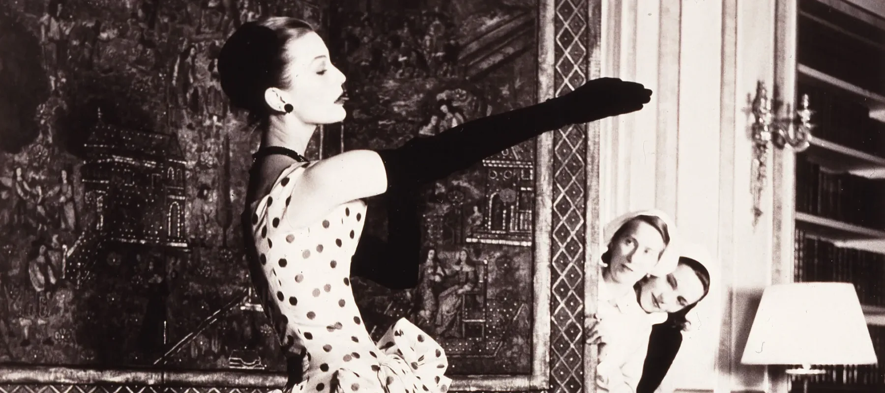 A black-and-white photograph of a woman with light skin tone in an elegant dress with dark polka dots and a bubble hem. The woman is putting on long black gloves while two women with light skin tone wearing uniforms peek around the corner of a large decorative panel to watch her.