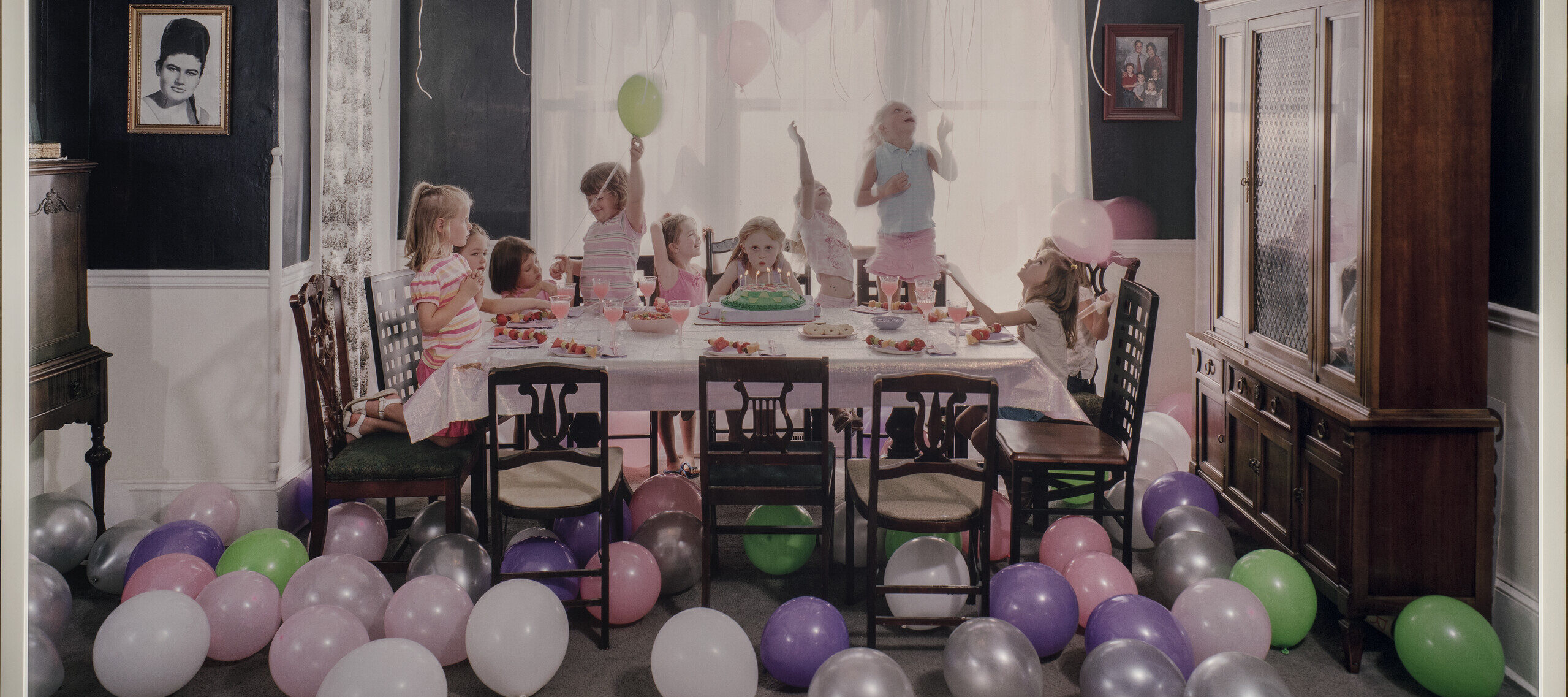 Green, purple, silver, white and pink balloons litter the floor and cover the ceiling of a navy-walled family dining room. In the middle of the room, a table is set with a pink table cloth. In the center of the table, a young girl blows out the candles on her birthday cake while looking straight ahead. Around her, nine other young girls jump, spin, and play.