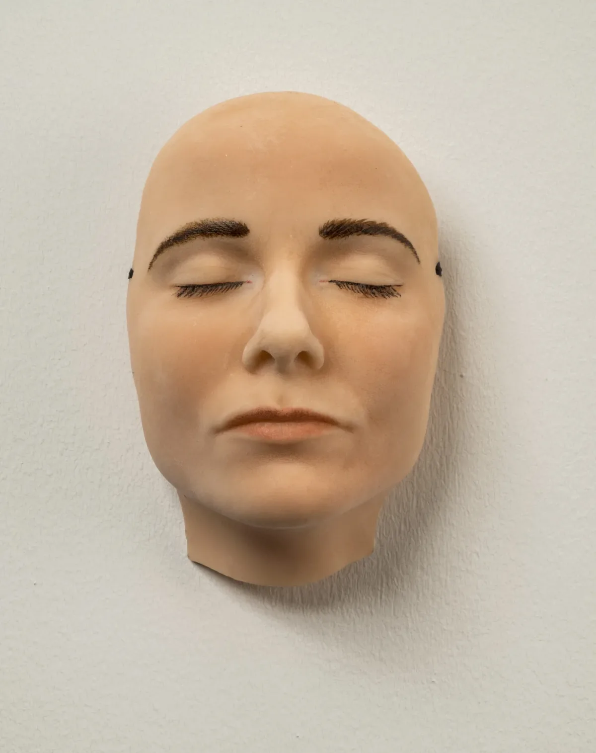 A highly realistic mask of a light-skinned face with dark brown eyebrows and eyelashes that appears to be sleeping—its eyes and mouth are closed and relaxed. The mask hangs on a white wall.