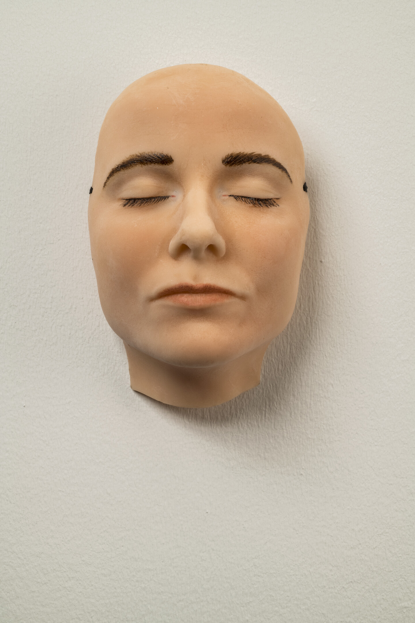 A highly realistic mask of a light-skinned face with dark brown eyebrows and eyelashes that appears to be sleeping—its eyes and mouth are closed and relaxed. The mask hangs on a white wall.