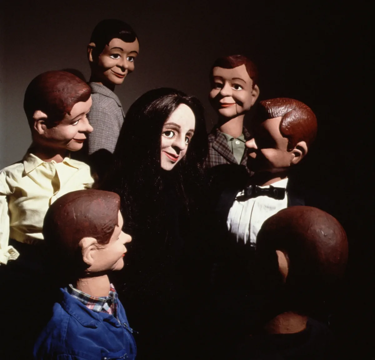 A light-skinned female ventriloquist dummy with straight black hair smiles in the center of a circle of six male dummy dolls with brown and dark hair.