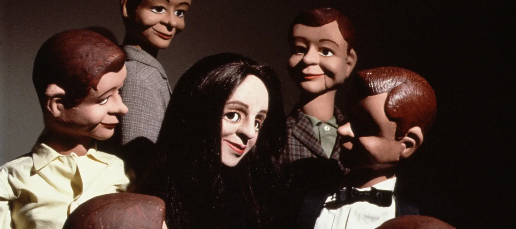 A light-skinned female ventriloquist dummy with straight black hair smiles in the center of a circle of six male dummy dolls with brown and dark hair.