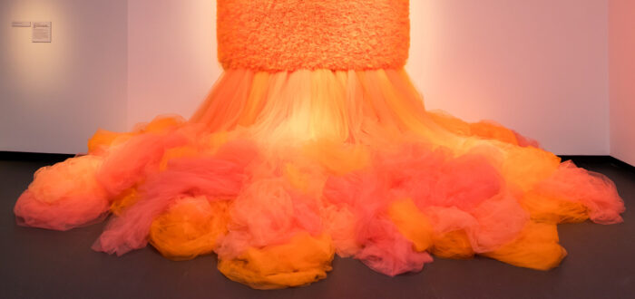 A work of art which is a large mass of orange tulle cascades down from a large, square tulle piece hanging on the wall.