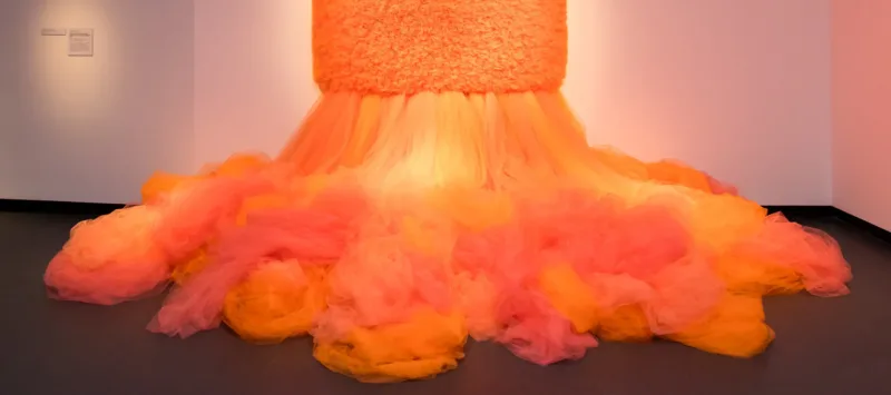 A work of art which is a large mass of orange tulle cascades down from a large, square tulle piece hanging on the wall.