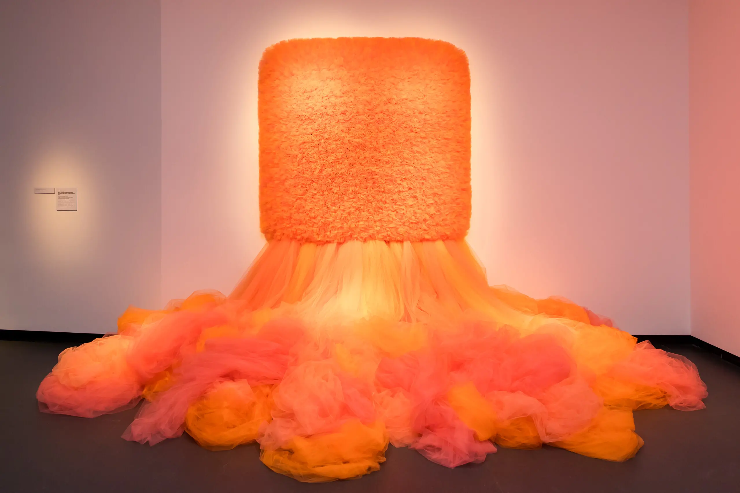 A work of art which is a large mass of orange tulle cascades down from a large, square tulle piece hanging on the wall.