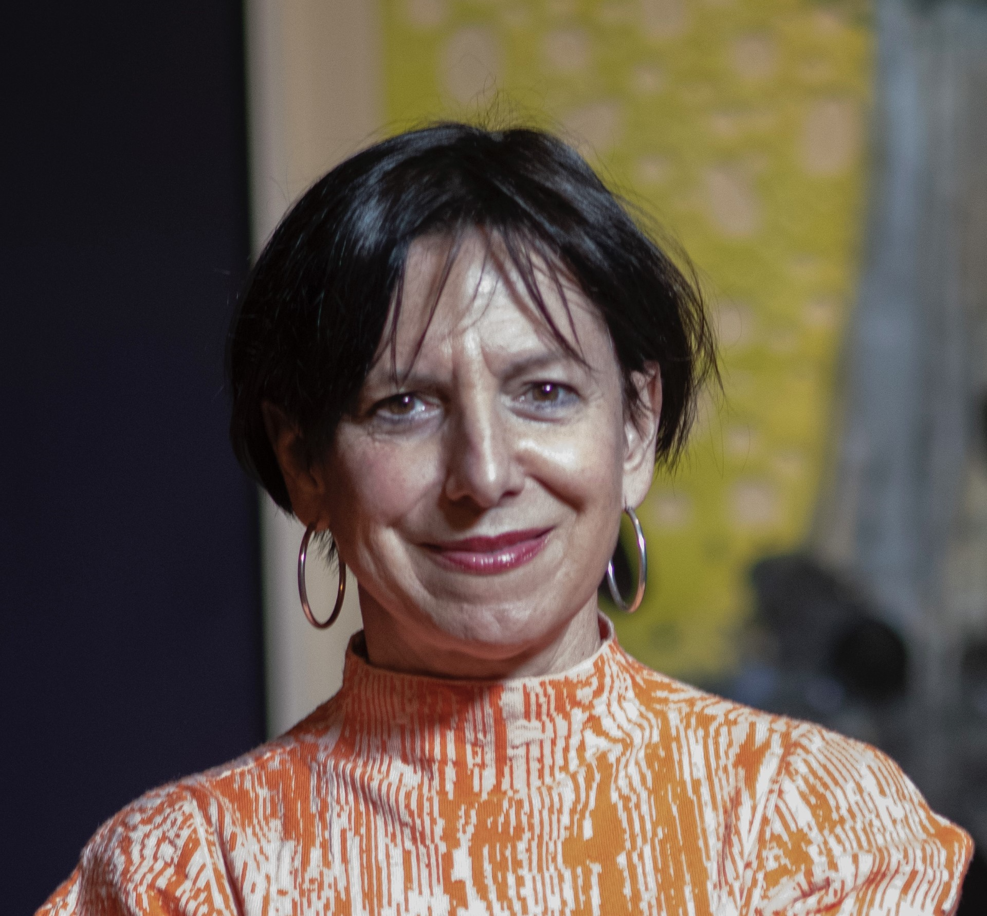 A light-skinned woman with short black hair smiles at the camera. She wears medium-sized hoop earrings and an orange-and-white graphic turtleneck.