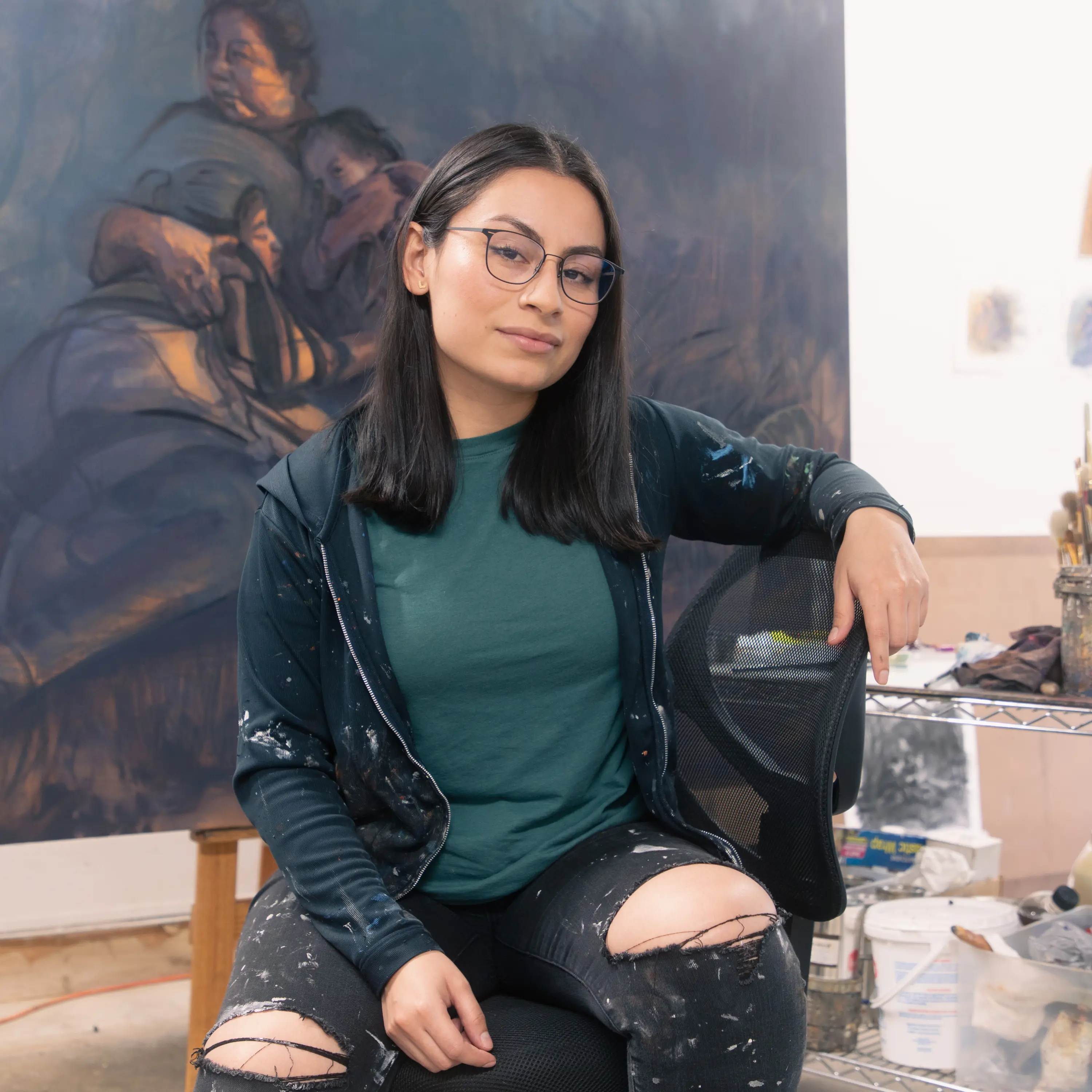 A young woman with Brown skin tone and straight black hair sits in front of a large painting. She wears a black hoodie and jeans that are peppered with paint and thin-rimmed black glasses. She sits in a chair and smiles slightly at the camera.