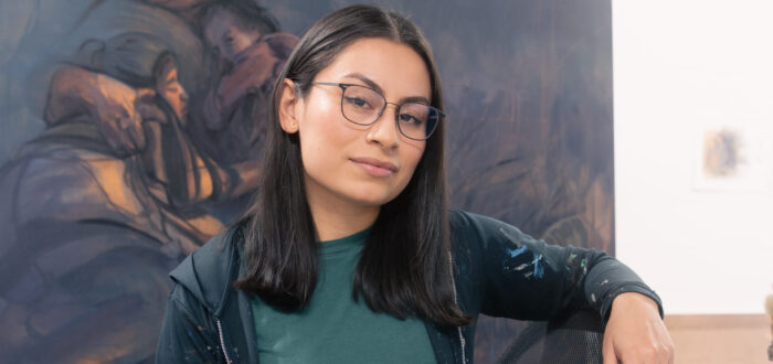 A young woman with Brown skin tone and straight black hair sits in front of a large painting. She wears a black hoodie and jeans that are peppered with paint and thin-rimmed black glasses. She sits in a chair and smiles slightly at the camera.