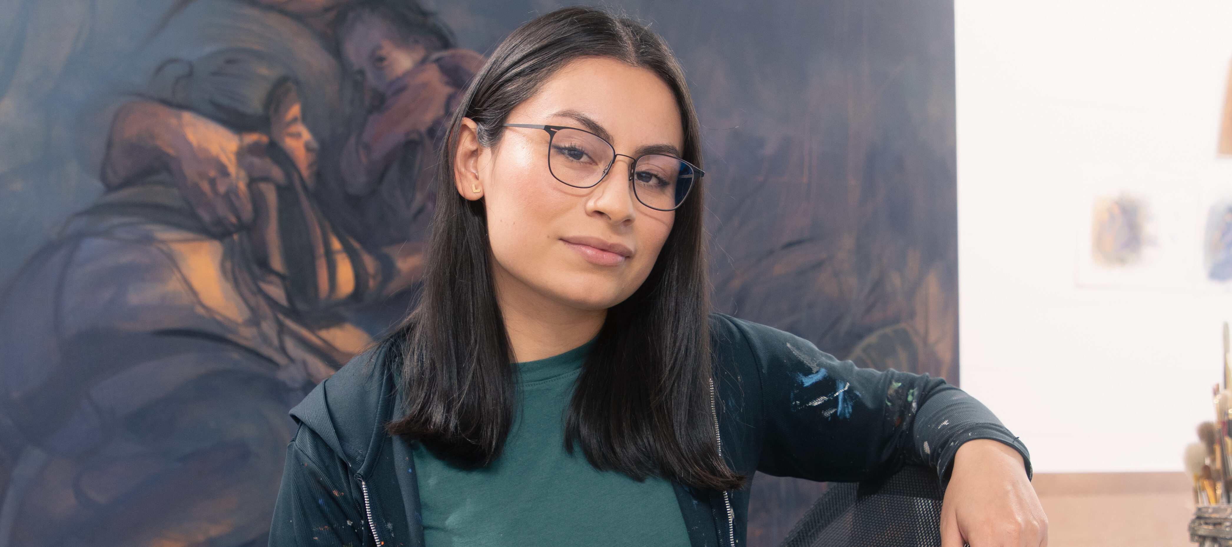 A young woman with Brown skin tone and straight black hair sits in front of a large painting. She wears a black hoodie and jeans that are peppered with paint and thin-rimmed black glasses. She sits in a chair and smiles slightly at the camera.