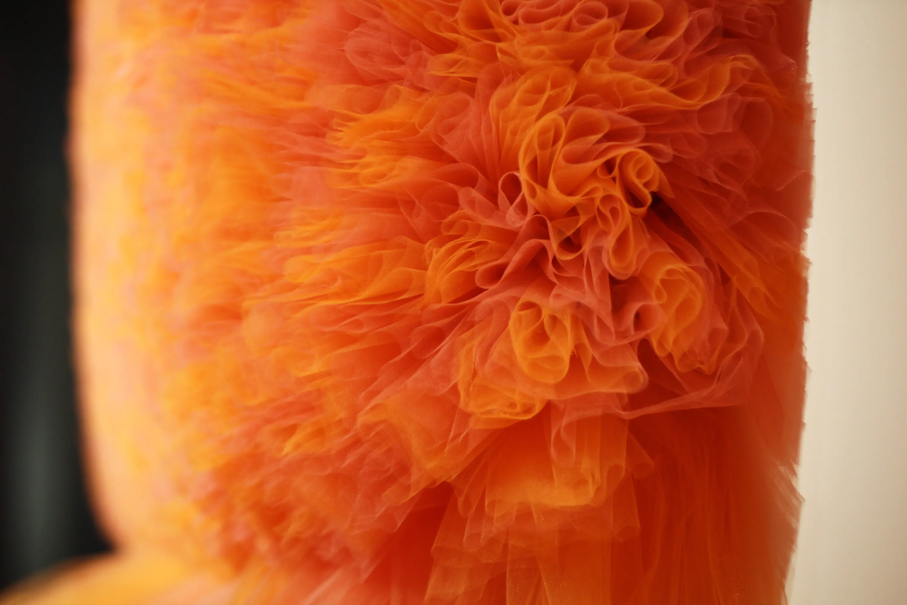 A close-up photograph of an abstract sculpture, made from bright orange and pink tulle, featuring layers and layers of the undulating fabric formed into a large circle.