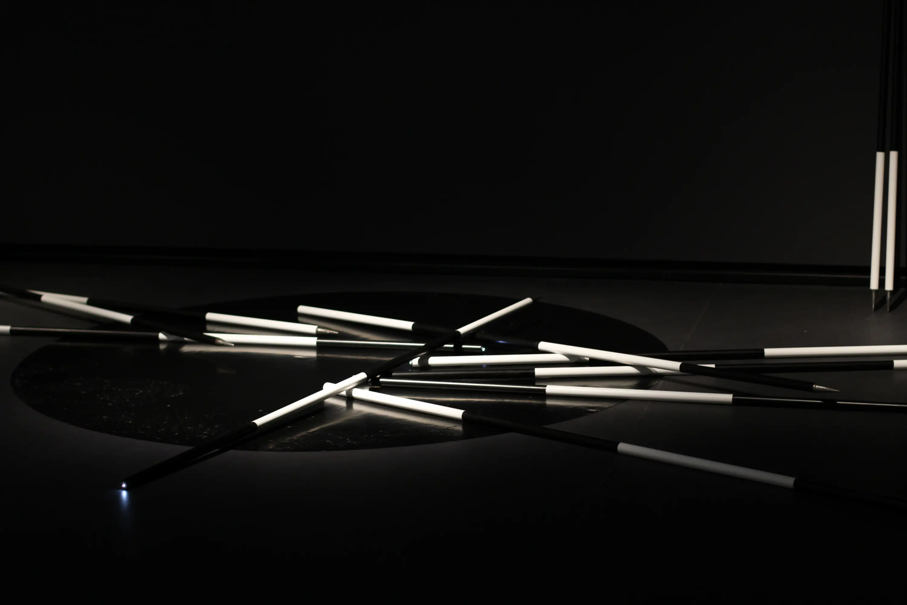 Black-and-white rods lay haphazardly over a black latex circle on the floor of a dark-lit gallery.