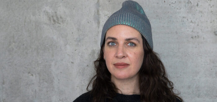 A light-skinned woman with long curly dark brown hair stands in front of a concrete wall. She wears a grey beanie and black shirt. Her light blue eyes are striking as she stares with a neutral expression at the camera.