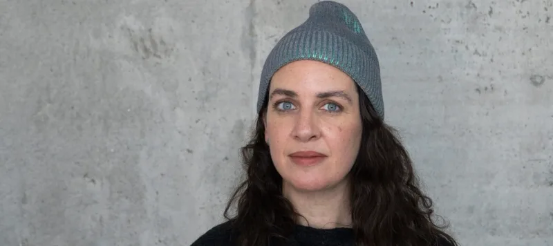 A light-skinned woman with long curly dark brown hair stands in front of a concrete wall. She wears a grey beanie and black shirt. Her light blue eyes are striking as she stares with a neutral expression at the camera.