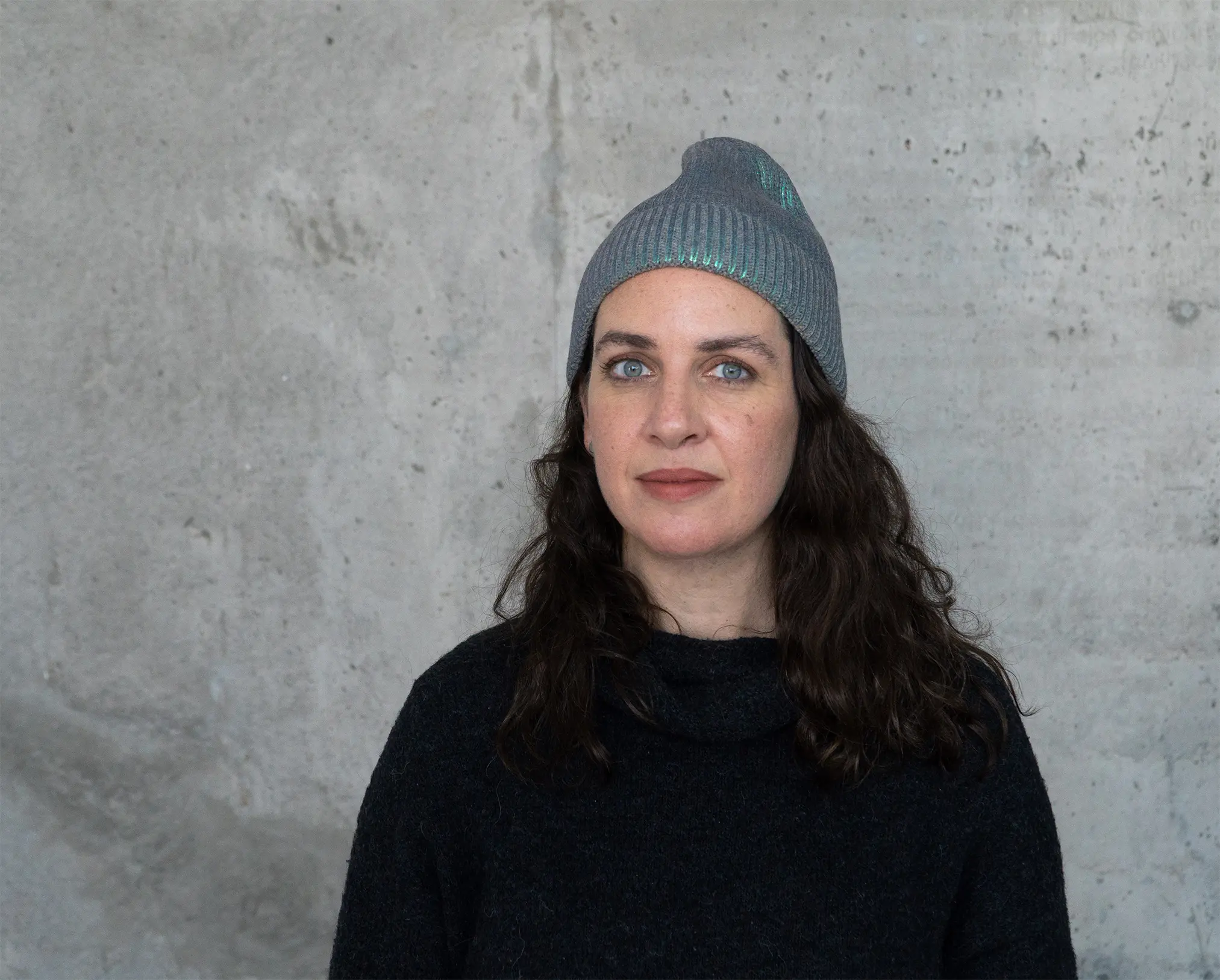 A light-skinned woman with long curly dark brown hair stands in front of a concrete wall. She wears a grey beanie and black shirt. Her light blue eyes are striking as she stares with a neutral expression at the camera.