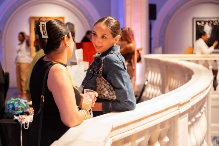 Two women look at each other, in conversation, holding drinks and leaning on a white marble railing.