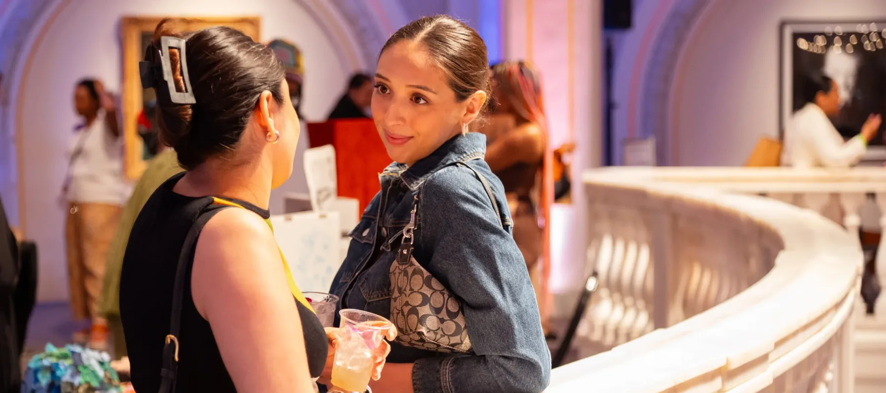 Two women look at each other, in conversation, holding drinks and leaning on a white marble railing.