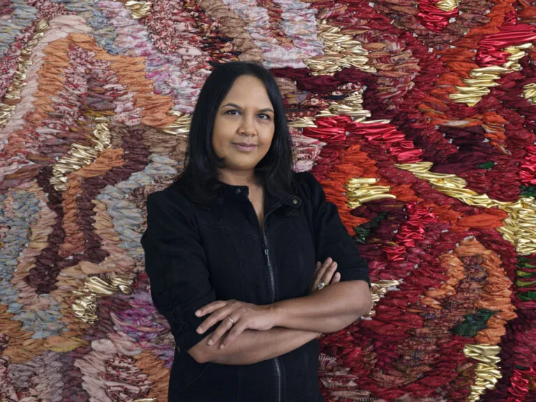 A photograph of a medium-skinned woman with black hair, shown from the waist up. She is wearing a black zip-up jacket and has her arms crossed in front of her. She stands in front of a large-scale abstract tapestry made of red, orange, pink, and gold fabric.