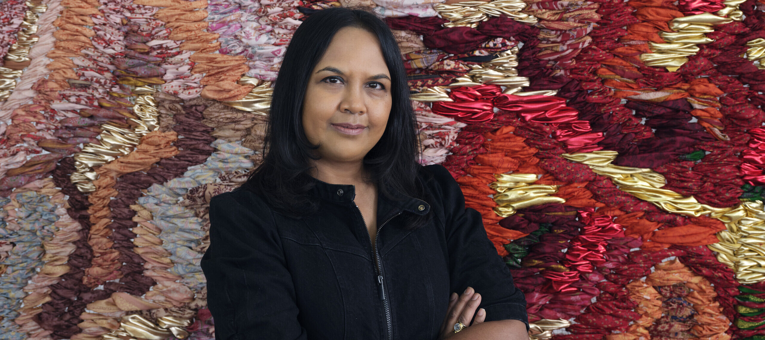 A photograph of a medium-skinned woman with black hair, shown from the waist up. She is wearing a black zip-up jacket and has her arms crossed in front of her. She stands in front of a large-scale abstract tapestry made of red, orange, pink, and gold fabric.