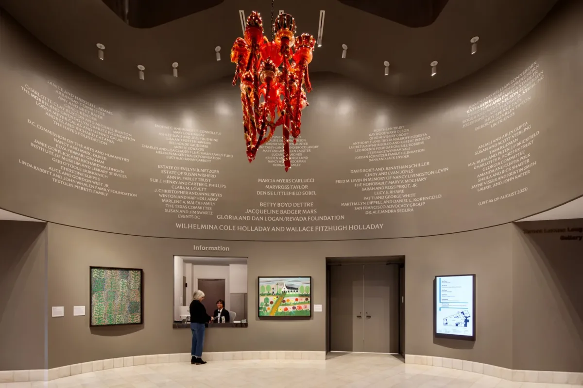 A museum rotunda features a red chandelier hanging from the center of the ceiling. The chandelier is made of red and orange glass and festooned with ornamented red-and-gold fabric garlands. LED lighting around the glass bulbs illuminates the chandelier.