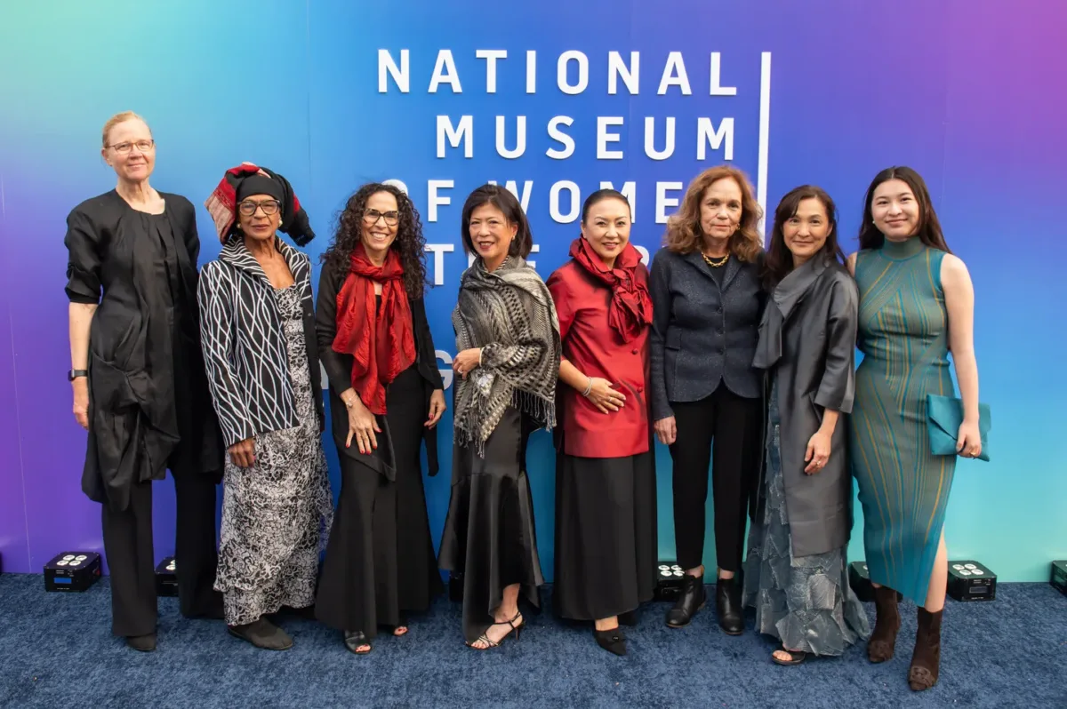 Eight women with various skin tones, all wearing evening wear, pose for a picture together in front of a backdrop that reads "National Museum of Women in the Arts Spring Gala 2024".