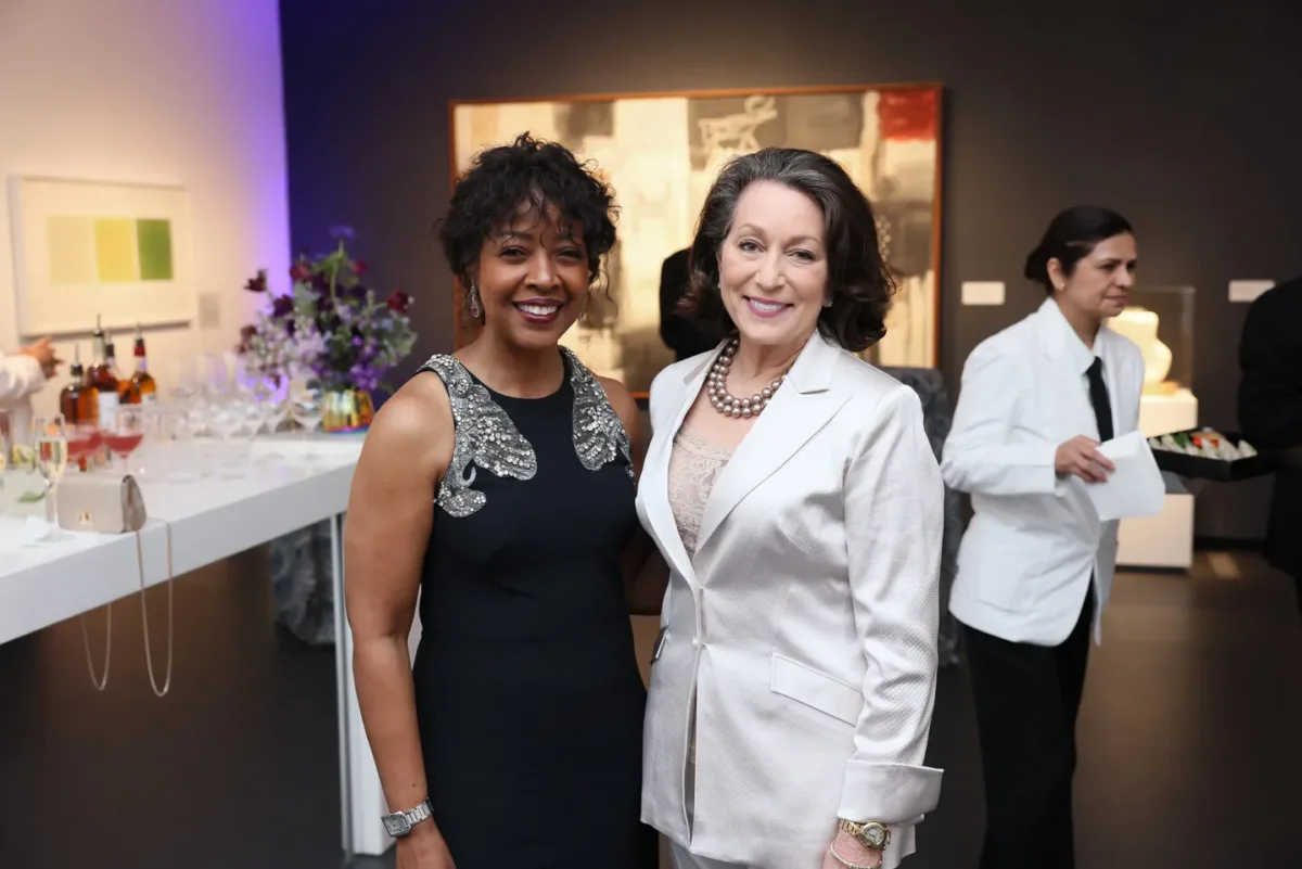 A woman with medium dark skin holding a black and silver evening dress poses for a picture inside an art gallery space with a woman with light skin wearing a white suit. Behind them, a waiter holds a tray of hors d'oeuvres.