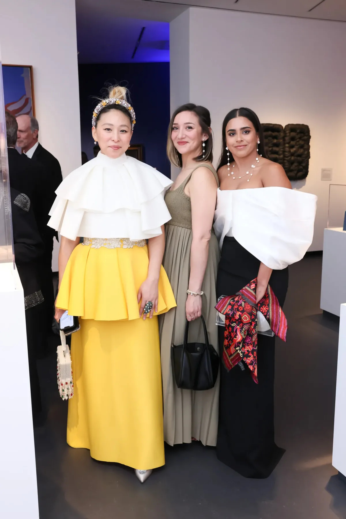 Three women with various skin tones wearing evening gowns pose for a picture inside of an art gallery space.