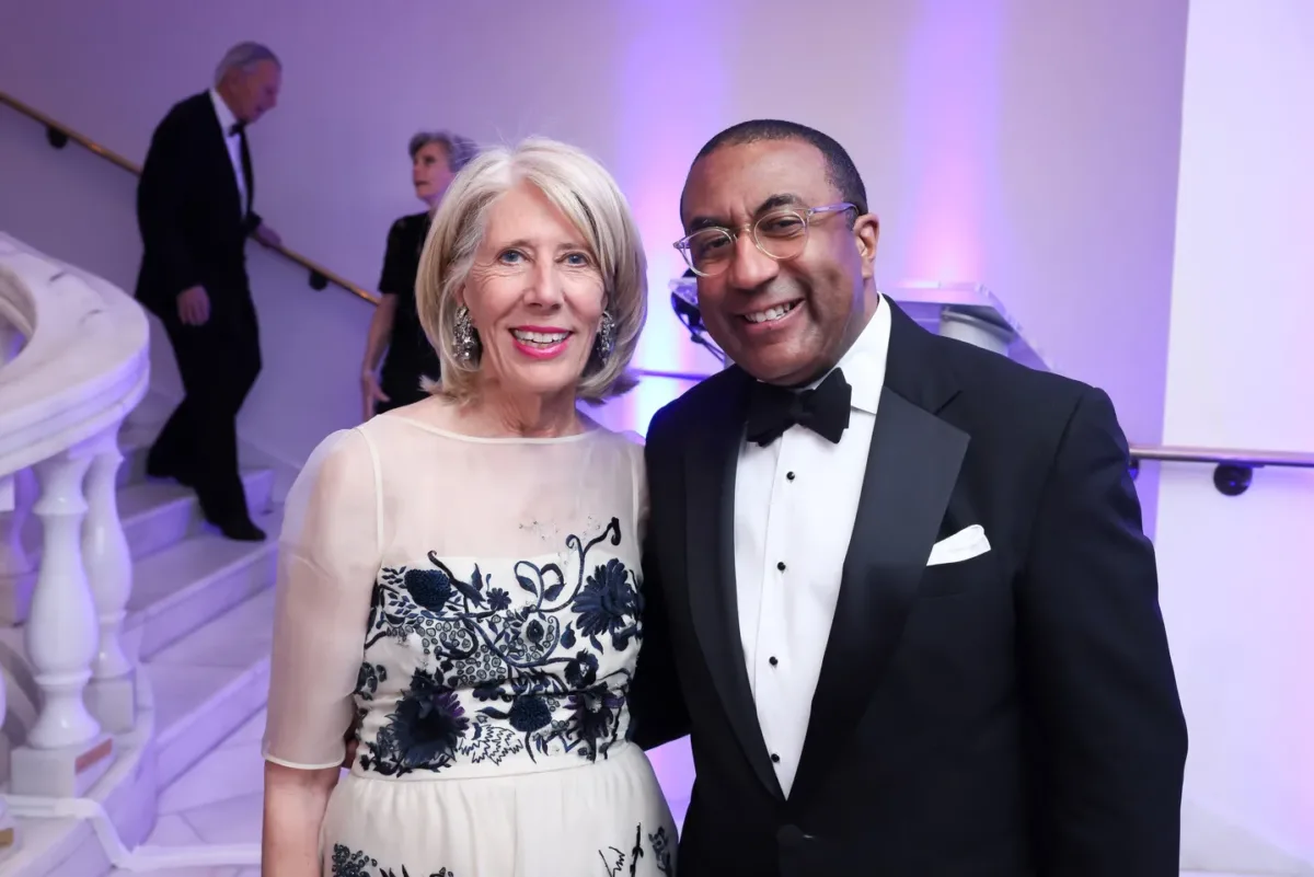 A woman with light skin and blonde hair wearing a blue and white evening dress poses for a picture with a man with dark skin wearing glasses and a tuxedo.