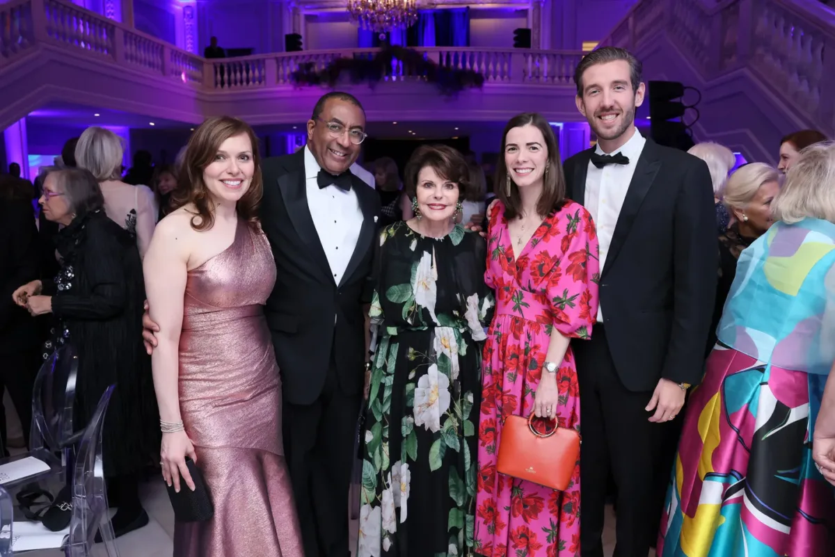 Three women and two men, all in evening wear, pose for a picture together inside of a large marble hall.