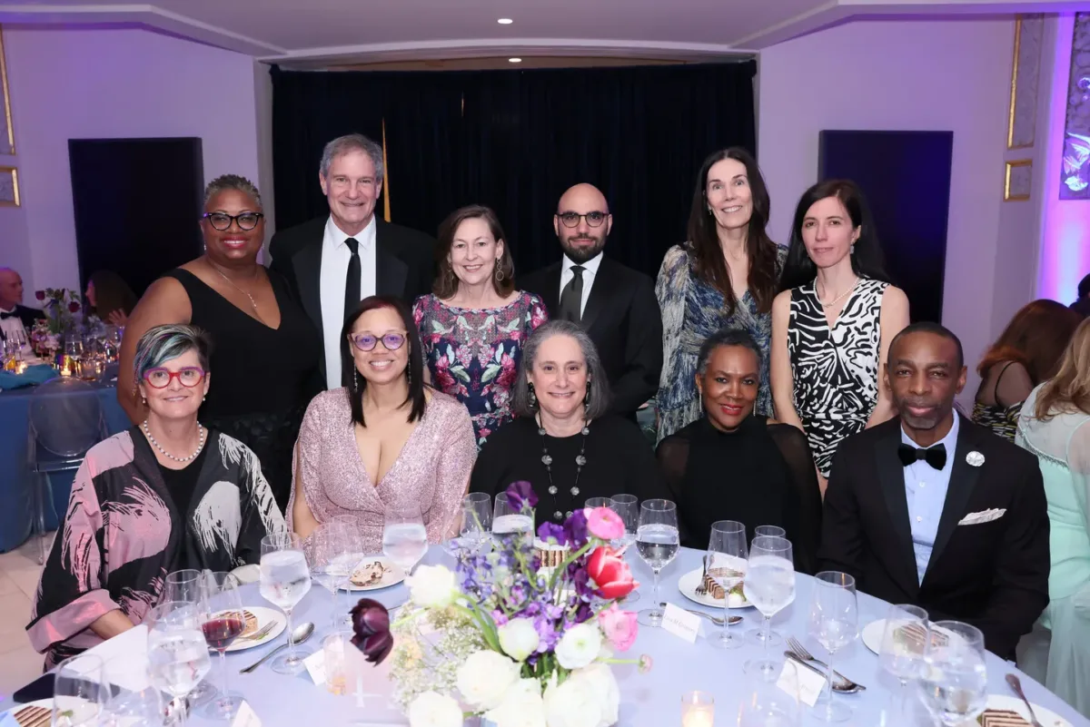 Eleven people, all in evening wear, pose for a picture together. Five people are seated while the remaining six people stand behind them.