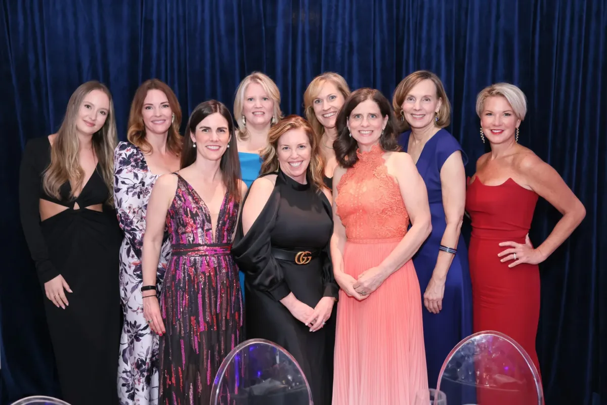 Nine women with light skin, all in evening wear, pose for a picture together in front of a navy blue backdrop curtain.