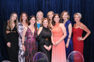 Nine women with light skin, all in evening wear, pose for a picture together in front of a navy blue backdrop curtain.