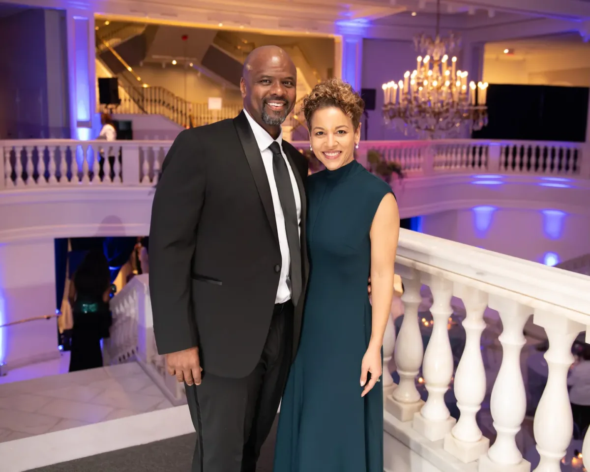 A man with dark skin wearing a black suit poses for a picture with a woman with medium skin wearing a dark green gown. They are at the top of  a white marble staircase.