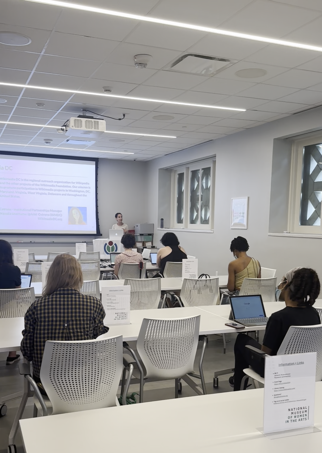 In a modern classroom, eight people sit at long white tables facing an instructor who stands at the head of the room next to a large projector.