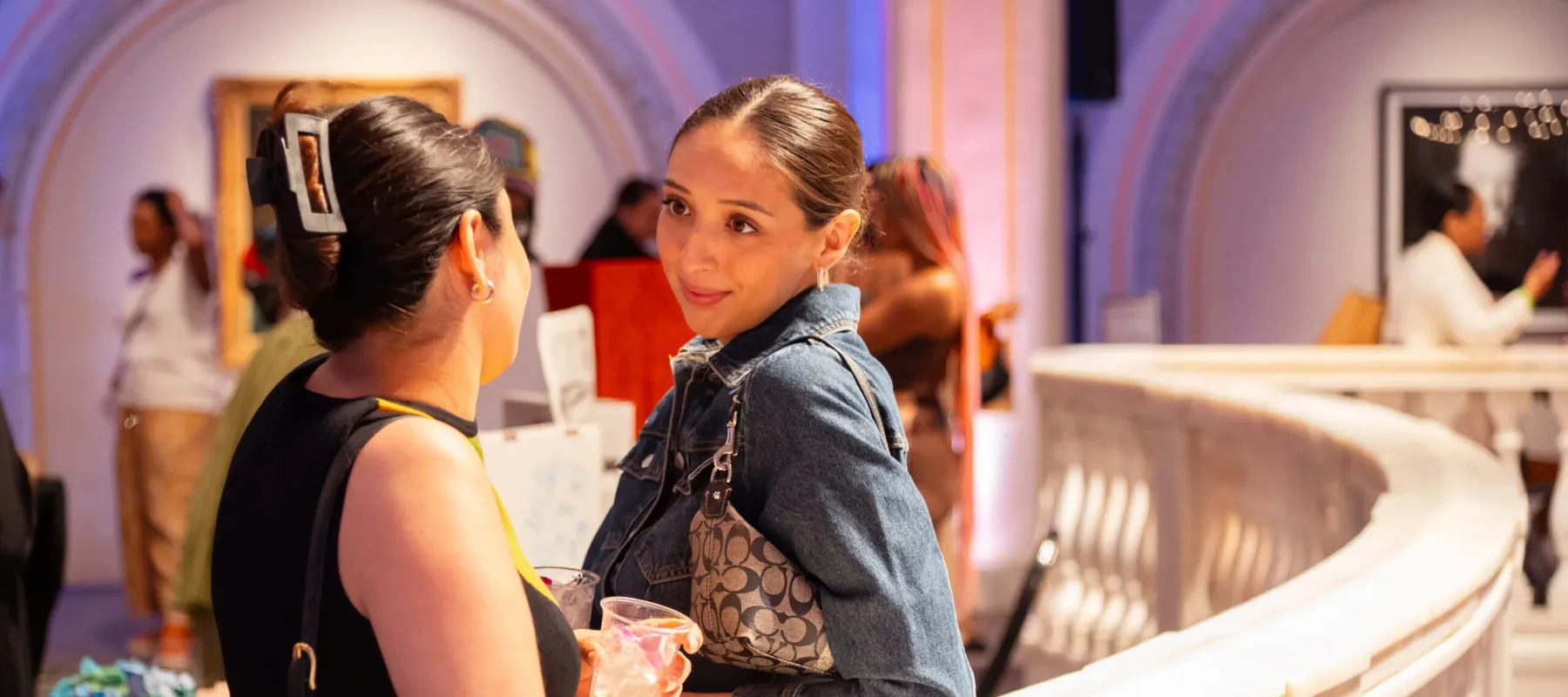 Two women look at each other, in conversation, holding drinks and leaning on a white marble railing.