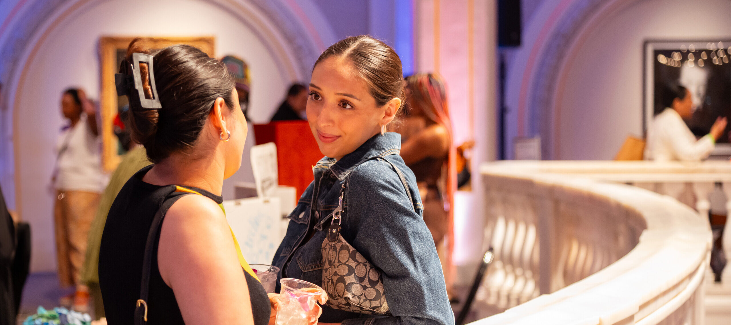 Two women look at each other, in conversation, holding drinks and leaning on a white marble railing.