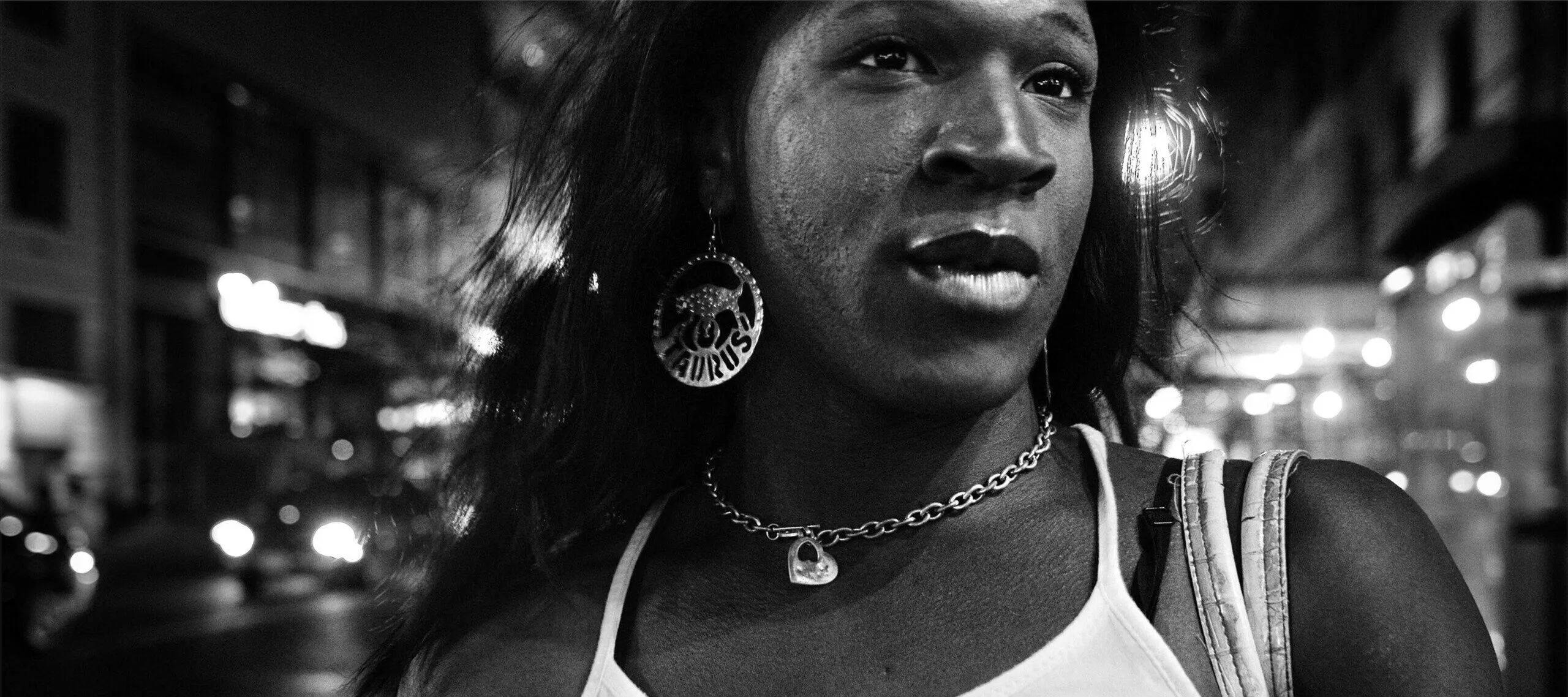 A black-and-white close-up image shows the face and upper body of a dark-skinned woman as she gazes off to the side. She wears a sleeveless white shirt, chain necklace with heart pendant, and large round earrings inscribed with the word “TAURUS.”